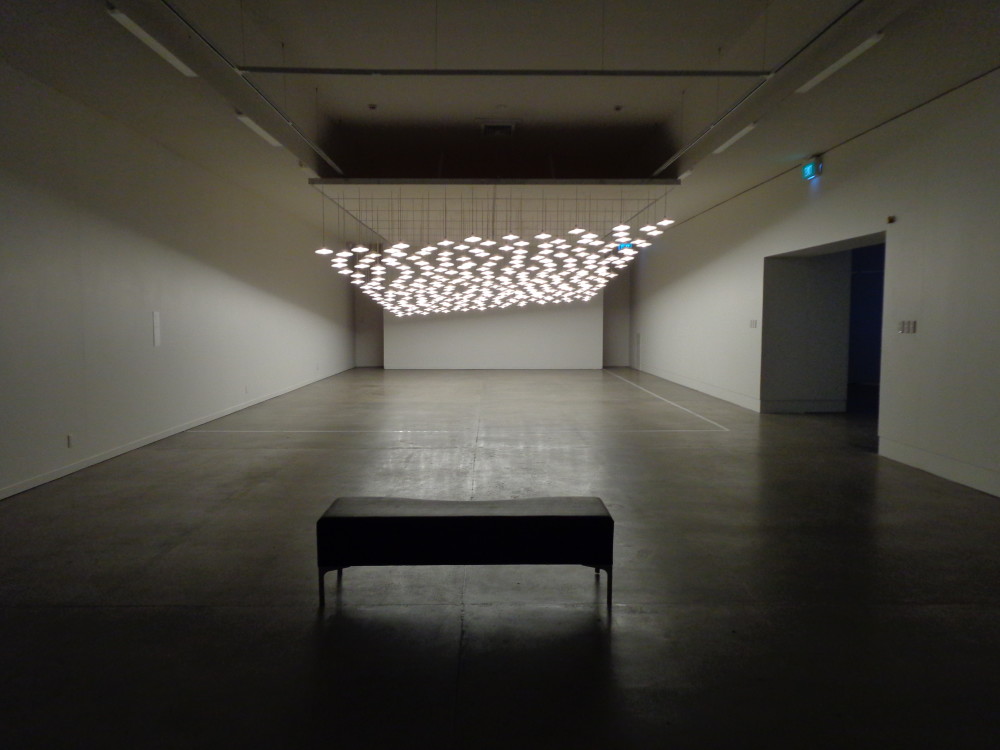 A long gallery corridor with a dark concrete floor, which has a single black bench placed in the centre foreground. At the other end of the corridor hangs masses of identical ceiling lights, which slant upwards at one end giving a tilted effect. They shine downwards and reflect light onto the floor below. 