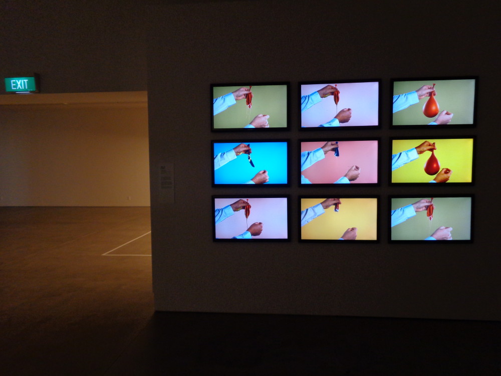Nine identically sized television screens are mounted in a grid on a darkened wall. Each screen has an almost identical still image of a person's hands popping a balloon. Each screen's video is slightly delayed so that two show the balloon still intact, and the others show it in various stages of popping. The background of each video is a different shade of pastel blue, pink, purple, orange or green. 