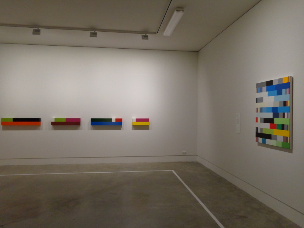 The image shows the corner of a gallery room. On the left is a series of giant cuisenaire rods stacked on top of one another, all in different colours, to form four individual oblong works. On the right is a visually similar artwork, a canvas painted with long colourful horizontal strips with white inbetween. 