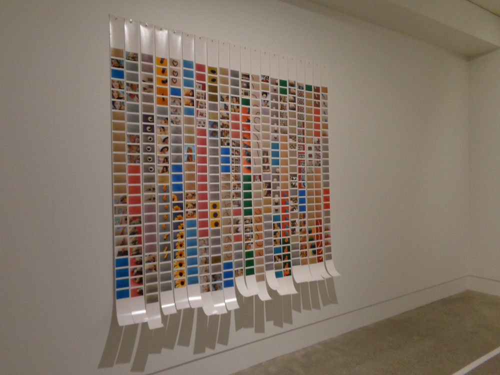 Multiple long strips of photographic glossy paper are pinned to a gallery wall, with their ends hanging loose and curling upwards. The strips have small photographs printed on them, with multiple photos being repeated in a line. Photographs include a close up of a sunflower, blue sky and people, but are too small to see clearly. The overall effect is of a colourful grid. 
