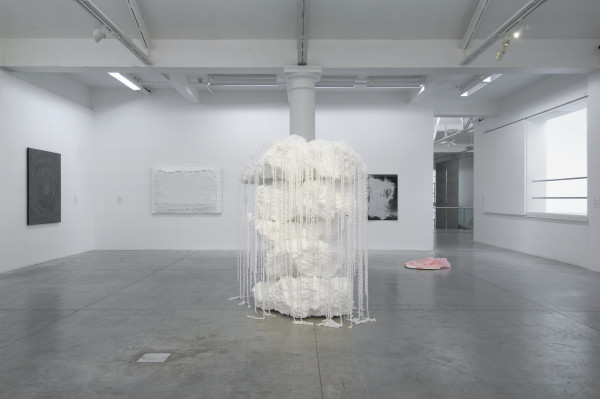 The image shows a tall stack of roughly cut polystyrene pieces, covered in thin white chains which are draped over the polystyrene stack and trail down to the floor. Behind this work three paintings are ivisible on a wall, and a piece of pink fabric is on a floor next to a corridor into another gallery room. 