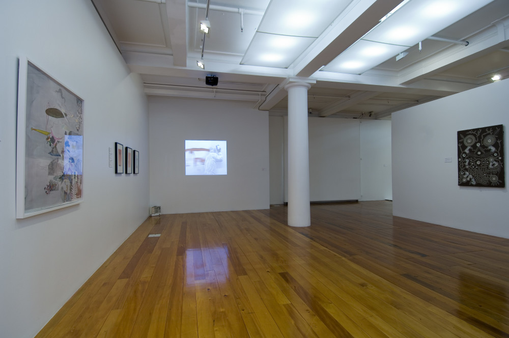 The image shows a white gallery space with a polished wooden floor. On the left hand wall is a large white watercolour in a white frame. On the back wall a video is projected onto the white wall, showing the artist in a white hazmat suit painting red strokes onto a wall using his head as the brush. The reflection from this work shines on the wooden floor beneath it. Next to the projection is a tall white floor to ceiling column, and on the far right wall hangs a brown painting with a series of circular patterns carved into it. 