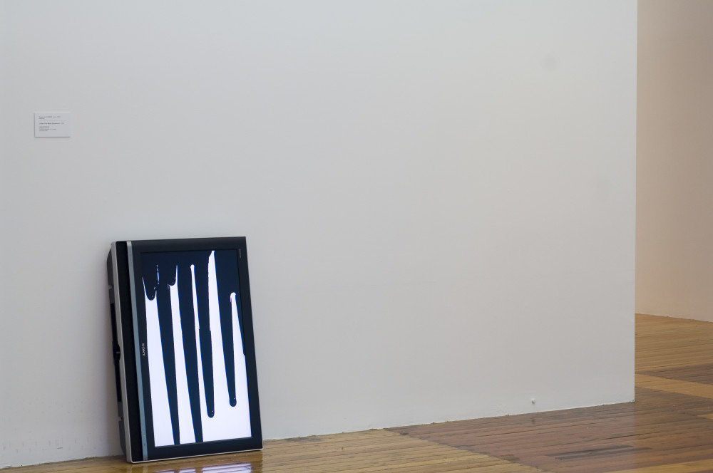 The image depicts a white gallery wall with a polished wooden floor below. A small TV screen is propped up vertically against the wall, with long lines of black paint appearing to drip down on the front of the screen over a white background. 