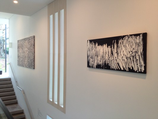 The image shows a gallery wall next to a staircase in Auckland Art Gallery. A vertical slatted window is in between two artworks. On the left is Bush Yam Dreaming, a work of grey circular spiralling shapes over a black background. On the right is a work by Dawn Naranatjil Yakuri, consisting of a black horizontal rectangular canvas with lots of long painted slashes of white paint filling the majority of the canvas. 