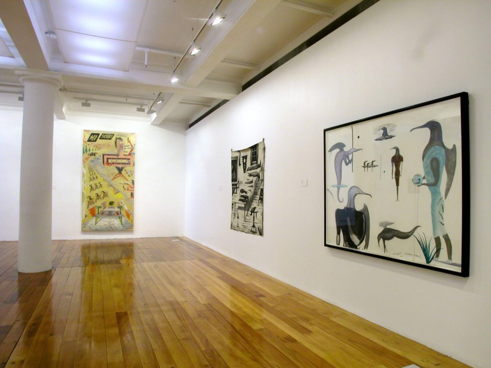 The image shows one half of a white gallery space with a polished wooden floor. A tall white column stretches from floor to ceiling on the far left of the image. To the right of this hangs a very tall vertical artwork, which depicts a composition with exaggerated perspective filled with imagery associated with radios, including antennas, a table of audio equipment, a car racing along a street and the words 'Hi Score Icon' in digitised lettering along the top of the artwork. All of this is on a mustard yellow background. On the right hand wall hang two paintings. On the left hangs a large square artwork which depicts a scene inside a home. An oblong table holds stylised birds of varying sizes and lengths stacked neatly in rows, while three more hang in a clump from the ceiling. Two taxidermied huia birds, with long beaks, are perched stiffly in a large glass vitrine to the left of the table. A glowing lamp and window looking out into a dark night are also present in the work. Across the bottom, the title of the painting, 'Buller's Tablecloth' is written in capital letters. To the right of this painting is a work filled with multiple, humanoid birds with long beaks and human arms and hands. Some have wings extending out of their backs, like angels. All are looking in different directions. 