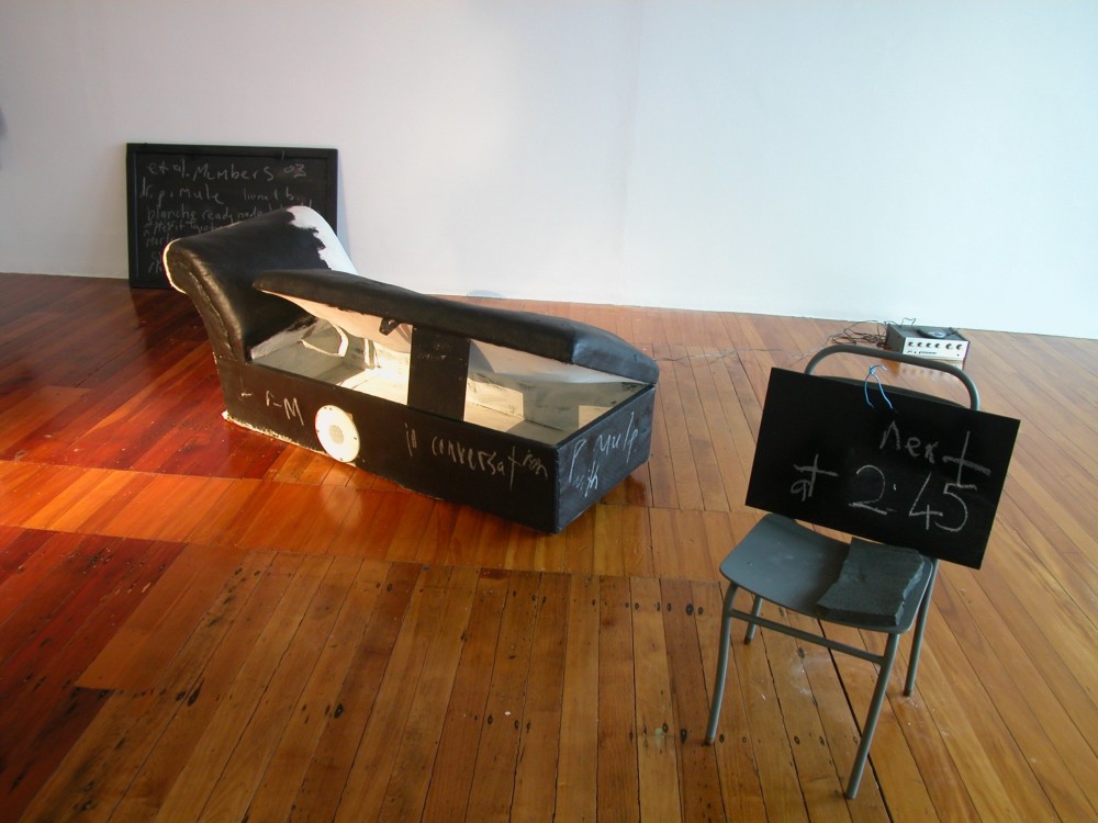The image shows a white gallery space with a polished wooden floor. A chalkboard with indistinguishable writing scrawled all over it is propped up against the back gallery wall. In the centre of the room a couch made out of black and white animal hide is located on the floor, with the seat opened and propped up by a small chalkboard to show the inside cavity of the couch. A white speaker is placed upon it. In the centre foreground, an old fashioned student's school chair, painted black, is positioned with another chalkboard propped up on it with 'next at 2:45' scrawled on it. An eraser sits on the seat of the chair.