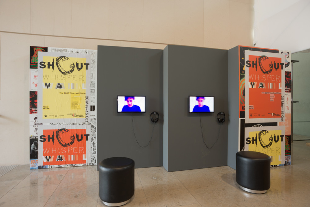 The image shows a large atrium space. Multiple freestanding temporary walls in grey are positioned in a staggered fashion. The two outside walls have promotional posters for the show on them in yellow and bright orange, while the two inner walls have small television screens mounted on them with headphones. A person in a blue shirt can be seen on the screens, taking up the whole frame so that their head and shoulders are the only parts of them visible. 