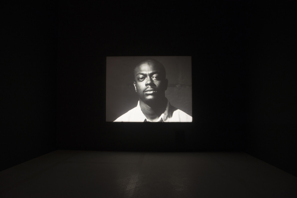 The image shows a large, dark room. In the centre of the room projected onto the back wall is a black and white image of a man with facial hair staring intently at the viewer, wearing a white shirt with collar. Only the head and shoulders of the man are visible, and he stands close to the camera taking up the frame. 