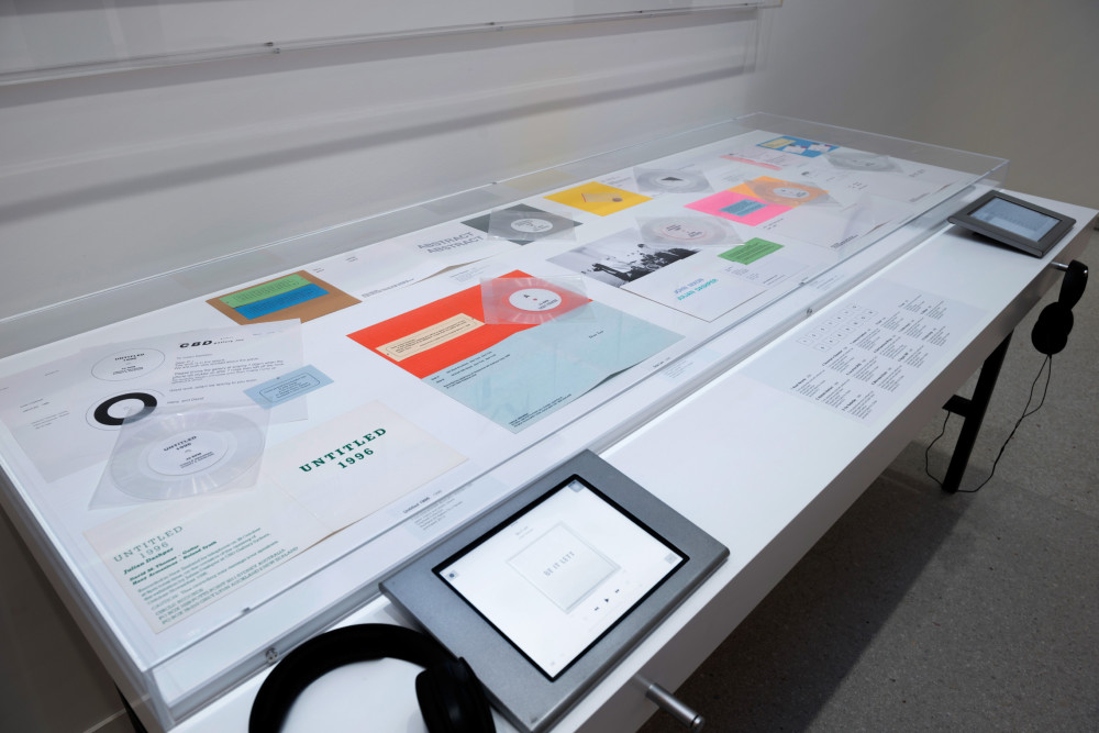The image shows a long, white vitrine table filled with ephemera. Items in the vitrine include vinyl records and covers in a variety of bright neon colours. In front of the table are two small interactive touchscreens and a pair of headphones. 