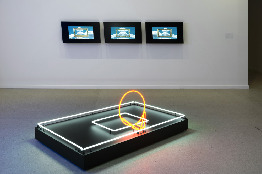 The image shows a gallery space. In the centre of the image on the floor is a large rectangular neon sculpture, comprised of a rectangular black platform outlined in white neon tubing, with a smaller rectangle in white neon in the middle. A circle of bright orange neon stands up at a 45 degree angle from this platform. On the wall behind this sculpture are three televisions, each with an image of a gold picture framed against a background of fluffy white clouds and blue sky. 