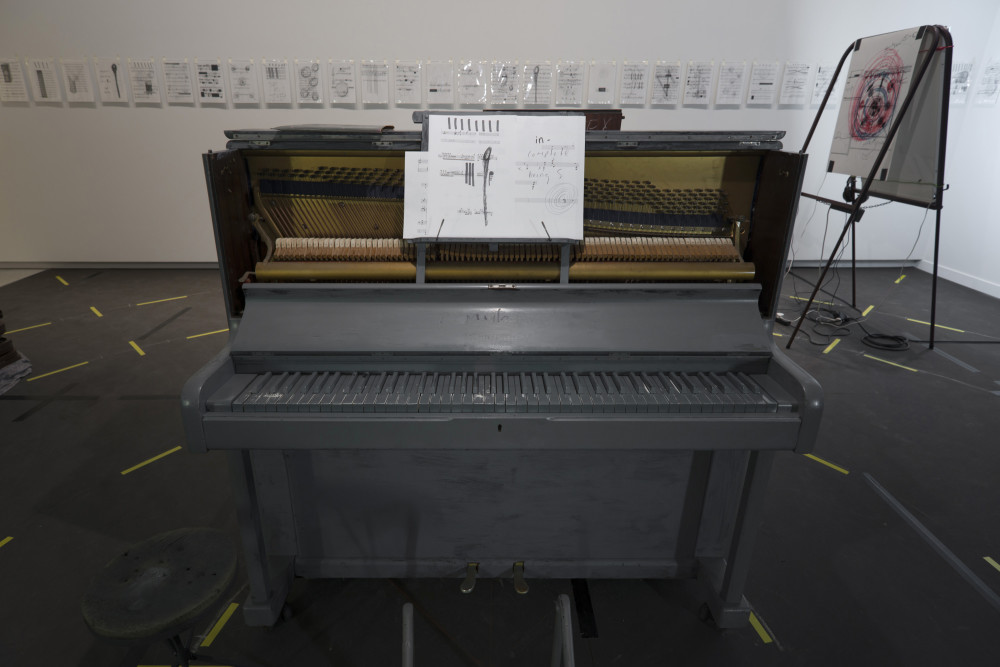 The image shows the same installation depicted in the previous image, but taken from the other side. In the centre of the image stands a large upright piano painted entirely in grey paint, with sheet music propped up on a stand above the keys. The sheet music is fragmented and has scrawls and scribbles all over it. Around the piano on the floor are the yellow off-cuts of tape circling the room, while on the wall behind the piano are multiple small drawings and prints in plastic protectors all in a row. To the right of the piano is one of the easels holding a large print. 