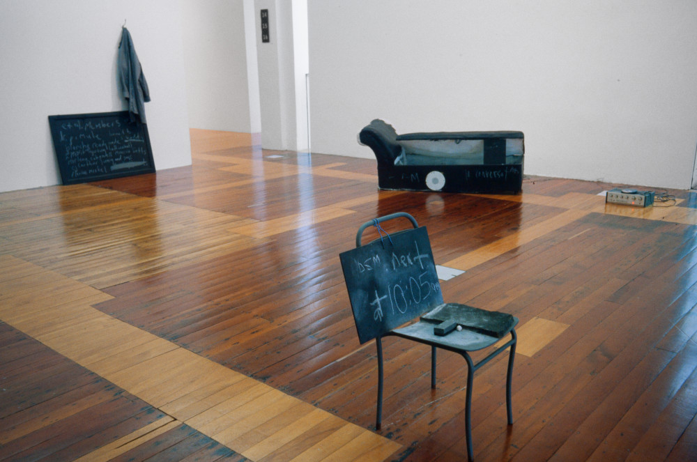 The image shows a white gallery space with a polished wooden floor. On the left white wall hangs a dark grey coat next to a chalkboard with indistinguishable writing scrawled all over it. In the centre background a couch made out of black and white animal hide is located on the floor, with the seat opened and propped up by a small chalkboard to show the inside cavity of the couch. A white speaker is placed upon it. In the centre foreground, an old fashioned student's school chair, painted black, is positioned with another chalkboard propped up on it with indistinguishable writing on it. An eraser sits on the seat of the chair. 