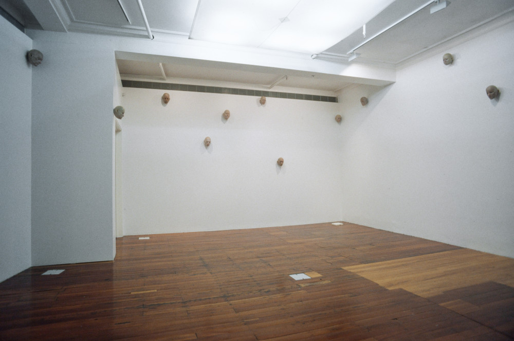 The image shows a white gallery space with a polished wooden floor. Hung across the walls in a scattered fashion are multiple small models of bald human heads, which look like mannequin heads. They are hung high up and are tilted down towards the viewer. 
