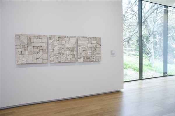 Three artworks called Web, Piece Work, and Foreign Affairs, all by Rosalie Gascoigne are shown hanging on a wall on the second floor of Auckland Art Gallery. The artworks are three square panels of the same size, all made up of irregular pieces of wood of slightly differing colours, in light ashy shades of off-white and brown. To the right of the artworks is a floor length window which looks onto Albert Park.