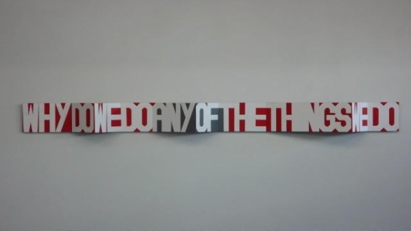 An artwork by Rose Nolan is shown displayed on a white wall. The artwork is a long strip of concertina folded card, with white lettering saying 'why do we do any of the things we do' taking up the whole strip of card. The spaces between the letters are filled with bright red and grey.