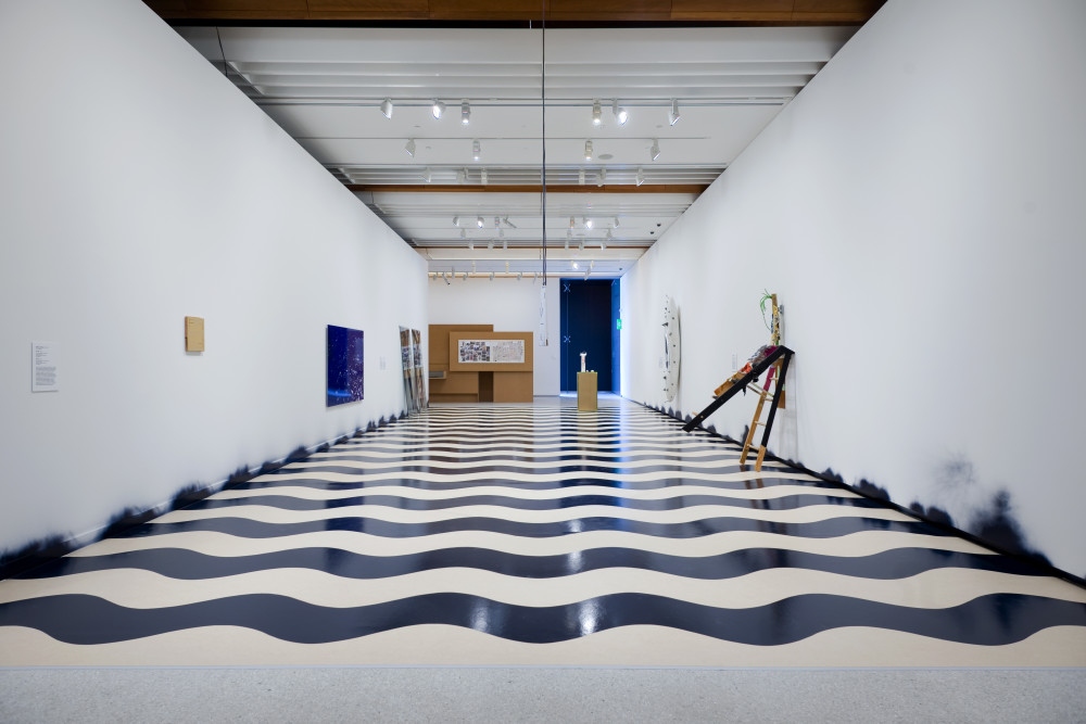 The image shows a long white corridor, looking from one end down to the other. On the floor of the corridor is a series of spray-painted black waves, which run horizontally down the length of the corridor. Black paint residue is smudged on the walls at the end of each of the waves. On the left hand wall is a small, light pink artwork hung high up from the floor, and further down is a large reflective dark blue square artwork. Another two tall sculptures are just visible leaning against the wall at the other end of the corridor. On the right wall, a white canoe hung vertically on the wall is just visible at the end of the corridor, and closer to the viewer is an easel opened out and leaning precariously against the gallery wall. At the end of the corridor a tall cardboard structure is visible but far away. 