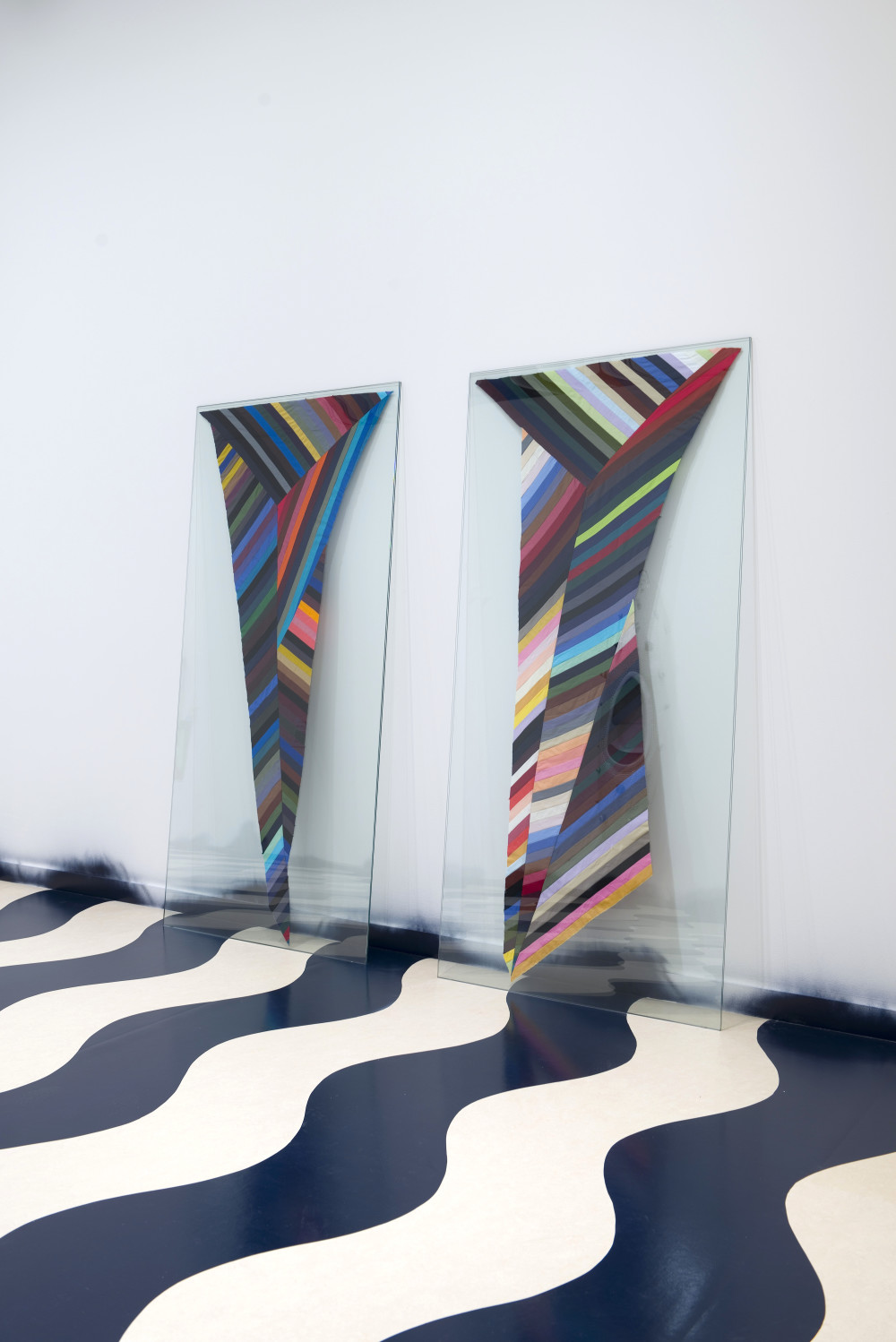 Two tall, vertical glass panes lean side by side against a white gallery wall. The floor below them is covered in a black and white wavy pattern. The glass panes have two similar but slightly different tapered designs sandwiched in between them, which are made up of thin strips of colour ranging from light pastel to dark blacks, including green, red, blue, purples, oranges, grey, and white in multiple shades. The strips are disjointed and formed trapezoid patterns within the design. The overall tapered effect in each of them is V-shaped. 