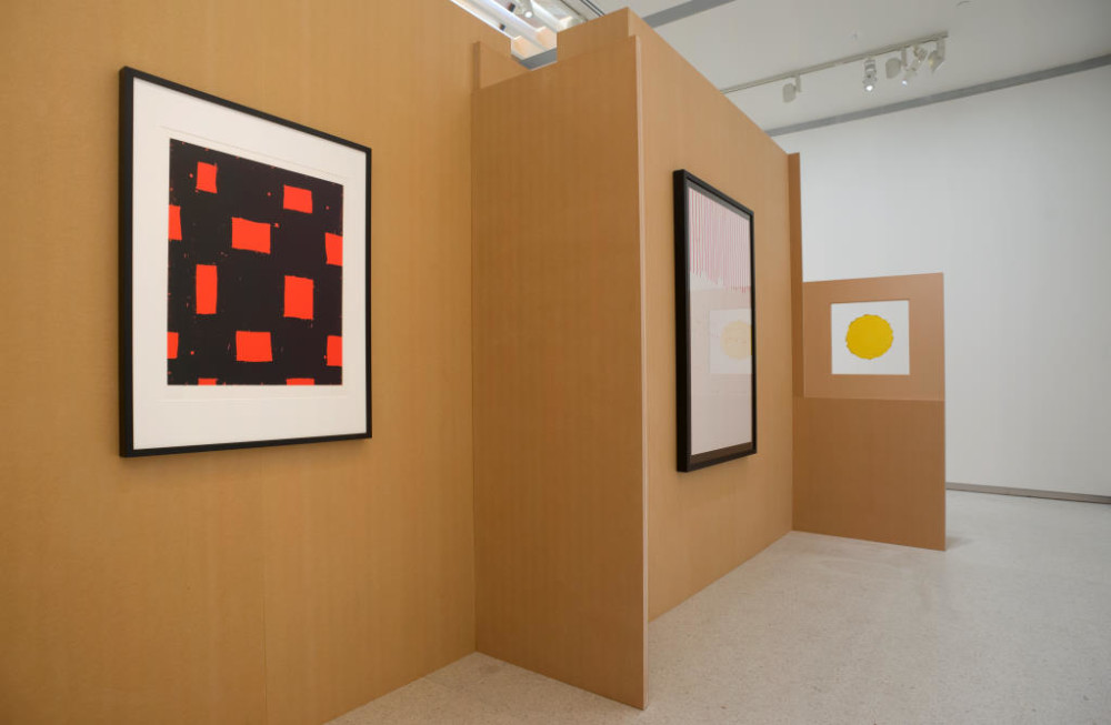 The image shows a closer view of the freestanding tall cardboard structure, which acts as its own bespoke gallery space. Mounted on the walls are three paintings: the first is a black screenprint with irregular oblong bright orange shapes scattered within it, the next has a white background with small vertical lines of red text running down it from the top of the print, and the third is a small white square with a round yellow circle centred in the middle. Each is contained in its own space next to one another. The entire structure sits within the main gallery space. 