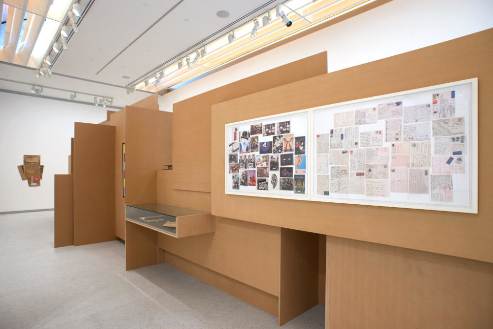 The image shows another part of the cardboard structure. On this side, another horizontal glass vitrine is visible in the left hand side of the structure, with two objects inside which aren't clearly visible. On the wall to the right of this vitrine hang two large framed collages of photographs and newspaper and magazine clippings arranged in a jumbled order. 