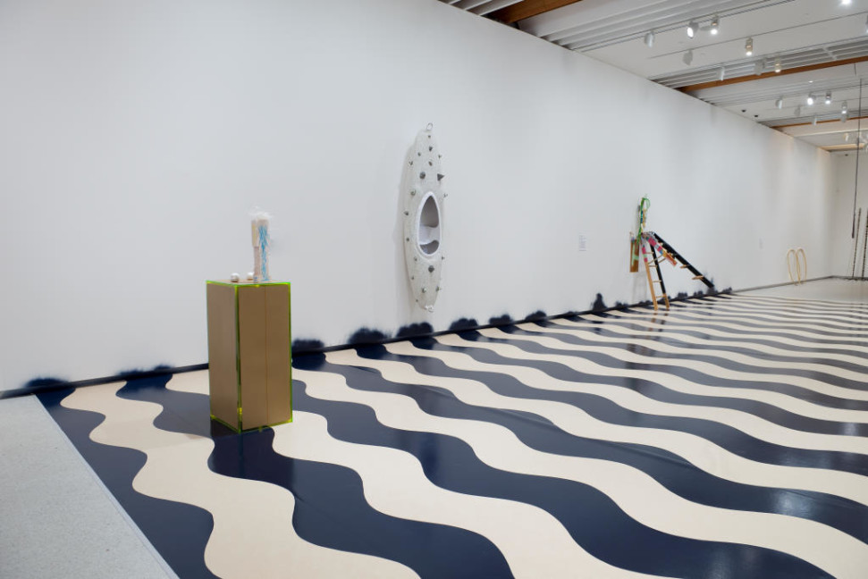 The image shows the corridor space from the other end of the room. The black and white waves ripple across the floor, and a tall narrow plinth edged in bright green with brown sides sits at the nearest end of the corridor with a smaller white painted figure perched on top of it. On the wall a little further down the corridor hangs a long white canoe flat on the wall, covered in large pieces of spiky dried paint which has been layered to produce a heavily 3D effect. These large masses are dotted all over the canoe hull. Further down the corridor, a small wooden stepladder leans against the wall, violently tilted so that its back legs are off the floor. 