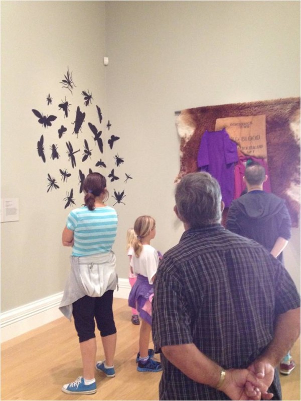 Gallery visitors (a woman, two children, and two men) stand in front of two artworks in a corner of Auckland Art Gallery. On the left hangs a circular arrangement of silhouettes of insects, and on the right wall hangs a large loose fur skin with bits of fabric sewn onto it.