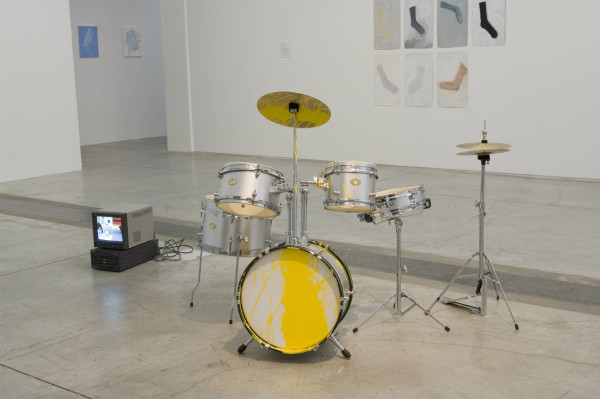 The image shows a junior sized drum kit in the centre of a gallery room, half covered in spilt bright yellow paint. To the left is a small cube monitor television screen, and in the background is a series of photographs of socks pinned to a white wall. 
