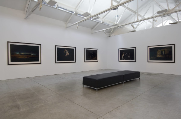 The image shows a room filled with a series of large photographs, side by side on gallery walls. The photographs are too dark to make out any detail in this image. A long narrow bench is placed in the centre of the room. 