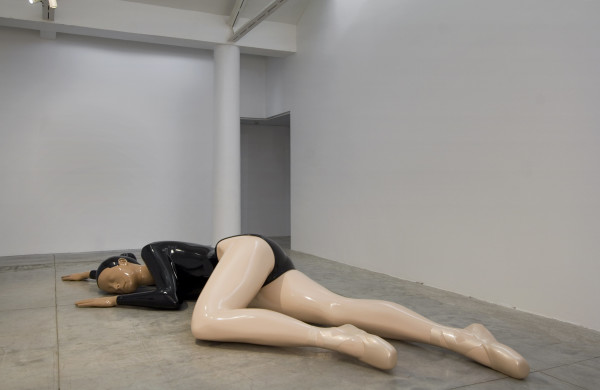 A giant sculpture of a ballet dancer is depicted in the image, lying down on the concrete floor on her side as if unconscious or asleep. She is wearing a long sleeved black leotard with light brown skin, pink tights and pointe shoes tied with ribbons around her ankles. Her black hair is arranged in a bun at the back of her head. The sculpture is extremely glossy and shiny due to the reflective automotive paint it has been covered in by the artist. 