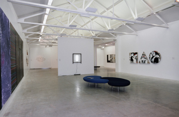The image shows a white gallery space with a concrete floor and high beam ceiling. On the left hand wall is a large, horizontal black and blue painting which is only just discernible to the viewer. On the centre back wall hangs a light work consisting of a vertical fluorescent tube inside a black frame. On the right hand wall hang three complimentary artworks, each in the shape of a square, triangle and circle, all painted with similar abstract enlarged brushstrokes in white, red and black paint against a gold background. 