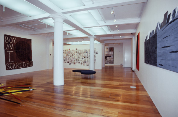 The image shows a white gallery space with a polished wooden floor. On the left hand wall hangs a large square artwork with the words 'boy am I scared eh' written in capital letters on it accompanied by a large black swirl. On the floor to the left of this work, half of a pile of giant pick-up sticks are visible. On the back wall hangs an assortment of smaller drawings closely jumbled together, each featuring a different small drawing including geometric patterns and humanoid shapes. On the right hand wall a diptych work of a dark blue and lighter blue panels are just visible. 