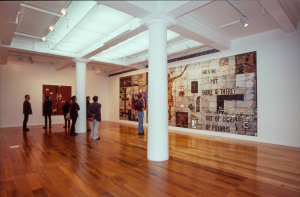 The image shows a white gallery space with a polished wooden floor. Several people mingle around in the space, looking at the artworks present. On the back left hand wall hangs a large square brown artwork with smaller scenes of miniature mountains dotted across its surface. On the right hand wall hangs a huge horizontal artwork made up of painted 'tiles', which features references to the work of Colin McCahon and Gordon Walters in its visual motifs, and has a large representation of Christ on the cross as its centre. 