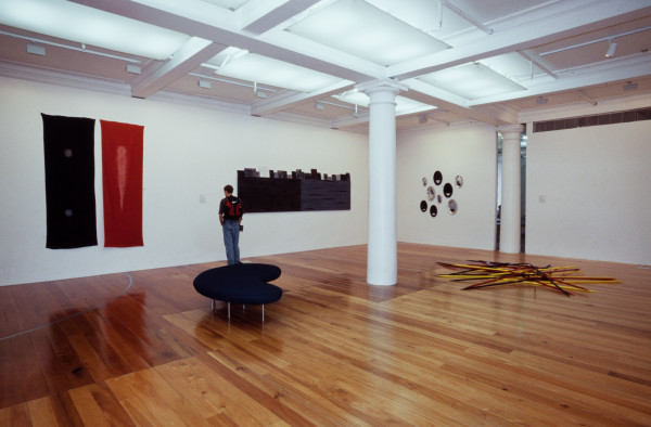 The image shows a white gallery space with a polished wooden floor. In the space, a middle aged man wearing jeans and a nike red and black backpack stands reading an object label. On his left hang two large vertical banners, one black and the other red. On the man's right is an artwork consisting of two large, horizontal panels, one in black and the other in dark grey. On top of these panels, multiple smaller works have been placed leaning against the wall facing the viewer. On the right hand gallery wall hangs an arrangement of metallic circles arranged in a circular fashion. On the floor to the right of this is a large installation of giant pick-up sticks. 