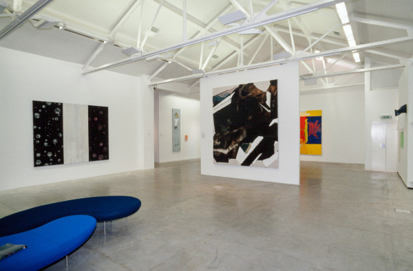 The image shows a white gallery space with a concrete floor and high beam ceilings. On the left hand wall hangs a horizontal artwork comprised of three panels, two in black with bubbles floating in them and the centre in white. On a central, freestanding wall hangs a large square artwork made from collaged photographs. In the background a bright multicoloured artwork is just visible. 
