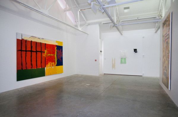 The image shows a white gallery space with a concrete floor. On the left hand wall hangs a large horizontal work filled with abstract squares and rectangles of bright colours, such as green, red, orange, blue and yellow. All are loosely painted and have a textured, messy effect. On the back wall hang some pastel coloured clothes, including a mint green skirt and white top. On the right hand wall a large horizontal painting is barely discernible. 