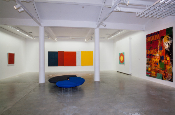 The image shows a white gallery space with a concrete floor. On the left hand wall hangs a small square artwork which is filled with a solid block of rust red paint, with a little texture below the surface. On the back wall hangs a large horizontal work made up of eight panels joined together at varying heights to give a wonky effect. Each panel is coloured in a solid block of dark blue, dark red, bright red, white and yellow. On the right hand wall at the back hangs a small square painting consisting of a yellow circle within a red circle within a bright blue background. Next to this, closest to the viewer is a large vertical artwork made up of overlapping squares and shapes of colour, with a large image of a young girl in black and white muddled into these. 