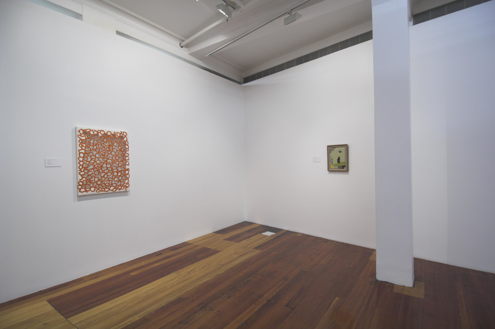 The image shows a white gallery space with a polished wooden floor. On the left hand wall is a rectangular abstract painting of red lines squiggled around on top of a grey background, with raw canvas edges. On the right hand wall is a much smaller, olive green painting showing a small black stylised figure standing on a hill, with a bird flying above and a small sapling to the left of the figure. On the far right of the image is a large white column which intrudes into the gallery space. 