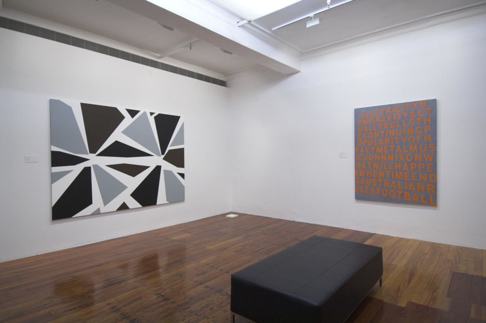 The image shows a white gallery space with a polished wooden floor. A low grey bench is placed in the centre of the space. On the left hand wall is a large landscape abstract painting depicting large shards of grey and black against a white background, clustering into two points. On the right hand wall is a large square painting depicting bright orange text on a dark grey background, which has a jarring contrast and makes the text difficult to read. 