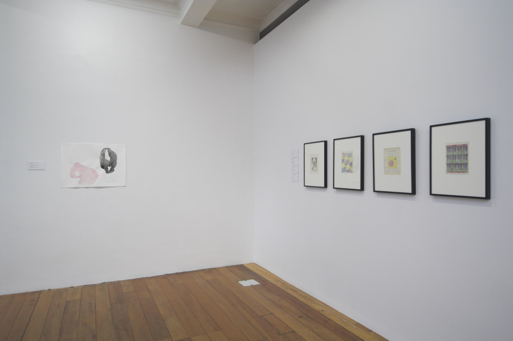 The image depicts the corner of a white gallery space, with a polished wooden floor. On the left hand wall is a small watercolour drawing of two pairs of underwear dropped on a floor, making abstract shapes. One is light pink, while the other is black. On the right hand wall is a series of black framed watercolours, of multicoloured geometric patterns. 