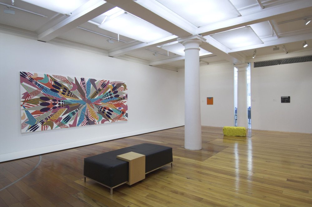 The image shows a white gallery space with a polished wooden floor. In the centre of the room is a low black bench. On the left wall is a large landscape painting showing a multitude of different multicoloured patterns all converging on a single point in the centre of the work, giving the overall effect of a mishmash of colours, shapes and overlapping patterns. On the right back wall of the gallery is a small rectangular abstract work showing an solid orange colour with a grey cutout point at the top of the frame. On the right hand side of this back wall is a small director's clapboard with 'The United States of Anxiety' scrawled on it in white chalk. In front of these two artworks is a large rectangular, yellow brick on the floor resembling a hay bale, but in an unnatural yellow colour. 