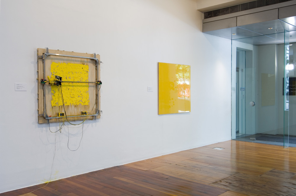 The image shows a white gallery space with a polished wooden floor. On the far right of the image, a set of sliding glass doors lead outside. On the wall next to these doors, on the far left hand side is an artwork consisting of a square sheet of plywood board, with a metal robotic contraption hooked up on it and in the process of painting a series of dabs of bright yellow paint onto the plywood. Next to this work is an orangey-yellow artwork, consisting of a large flat sheet of reflective metal with a spirit level running down one length of the work. 
