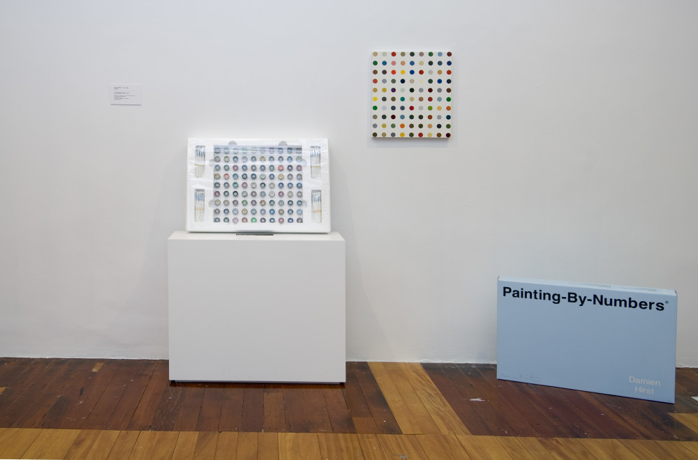 The image shows a white wall with a polished wooden floor below. On a plinth, leaning against the wall is a wrapped box of small individual paint pots in different colours, arranged in a grid pattern. On the wall next to this plinth hangs a small square painting covered in multicoloured, uniform dots which echo the pattern and shape of the paint pots in the box next to it. On the ground next to this is the light blue lid of the box, which has 'Painting By Numbers' written on it in black lettering, and the artist's name in white in the lower corner of the lid. 