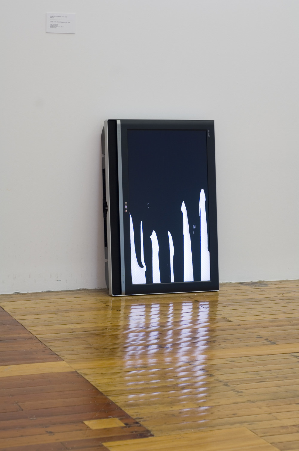 The image shows a television resting vertically against a white gallery wall and a polished wooden floor. Black paint appears to drip further and further down the white screen until it almost covers it. The reflection of the screen can be seen in the floor below. 