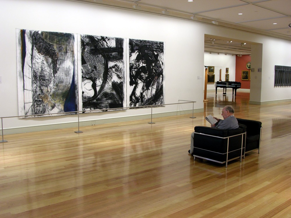 The image shows a white gallery space with a polished wooden floor. On the left hand wall hangs three floor to ceiling paintings made up of similar black patterns on white backgrounds, of varying textures and shapes. An older man sits in a chair in the centre of the room facing the painting, reading a piece of paper. Through a doorway next to the painting, more gallery spaces filled with sculptures and art on the walls can be seen. 