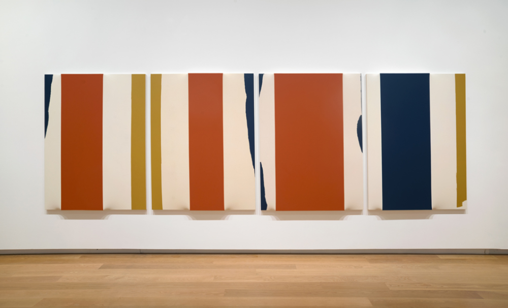 A long artwork made up of four canvases hangs on the wall in the gallery space. The canvases are covered with thick vertical stripes of brown, beige, mustard yellow and navy blue. The canvases bulge out at random points, creating a distorted effect.