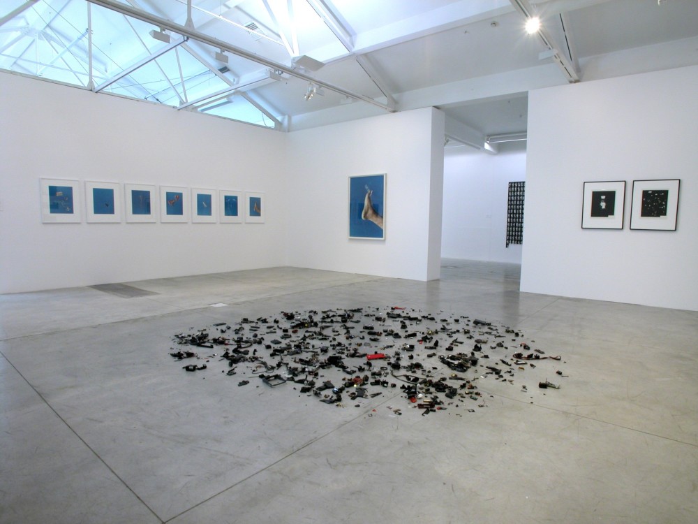 The image shows the other side of the gallery room which was shown in the previous photo. In this image, the same installation of miscellaneous mechanical pieces is visible scattered on the floor. On the far wall of the room, a series of seven visually similar photographs are just visible, all with white frames and light blue backgrounds. On the wall to the right of these is a large framed photograph of a person's foot, outstretched against a blue sky. To the right of this is a tall doorway leading into the next gallery space, and to the right of the door two smaller black framed photographs hang on the wall. 