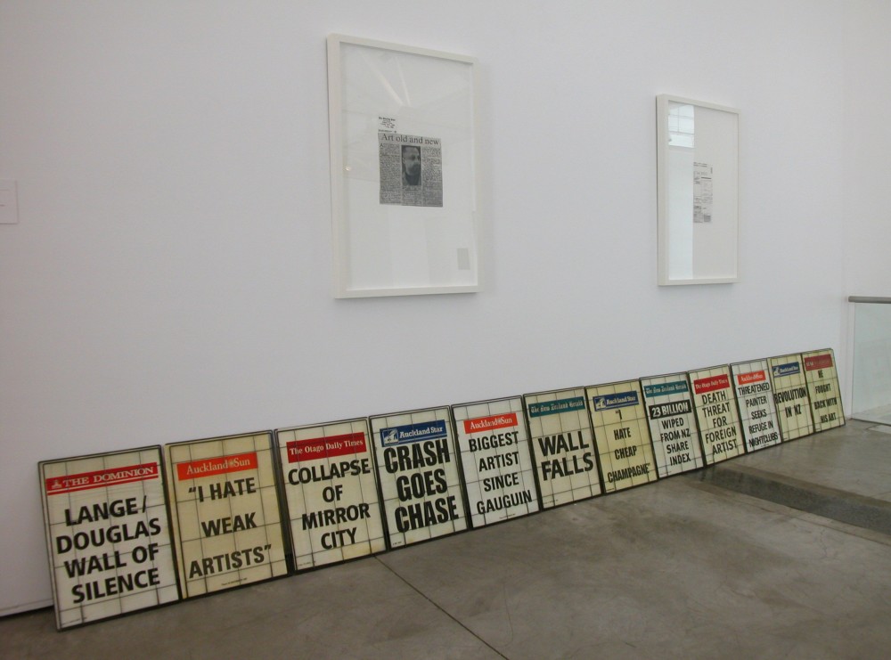 The image shows a white gallery wall and a concrete floor. On the floor a long row of newspaper headlines lean against the gallery wall, depicting a range of headlines contemporary to the time: including 'Lange/Douglas wall of silence', 'Wall Falls', 'Collapse of Mirror City' and Biggest Artist Since Gauguin'. Above these newspapers hang two newspaper clippings framed in large white frames. 