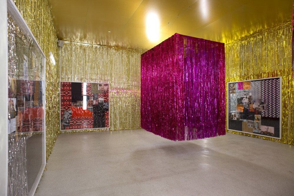 The image shows a rectangular tall gallery space. The walls of the gallery are covered in long strands of gold tinsel, from ceiling to floor. On each of these walls hangs a large white frame filled with collaged photographs of people, landscapes and patterns cut from magazines. In the centre of the room a large rectangular curtain of bright pink tinsel is suspended from the ceiling, floating like a large glitzy rectangular structure with a three dimensional appearance. 