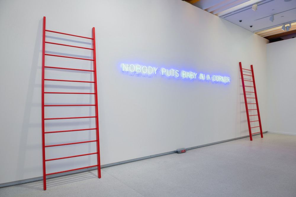 The image shows a wall in a gallery space. Leaning against the wall are two oversized wide red ladders. In between these ladders is some blue neon text mounted on the wall, which reads in capital letters 'No one puts baby in a corner'. 