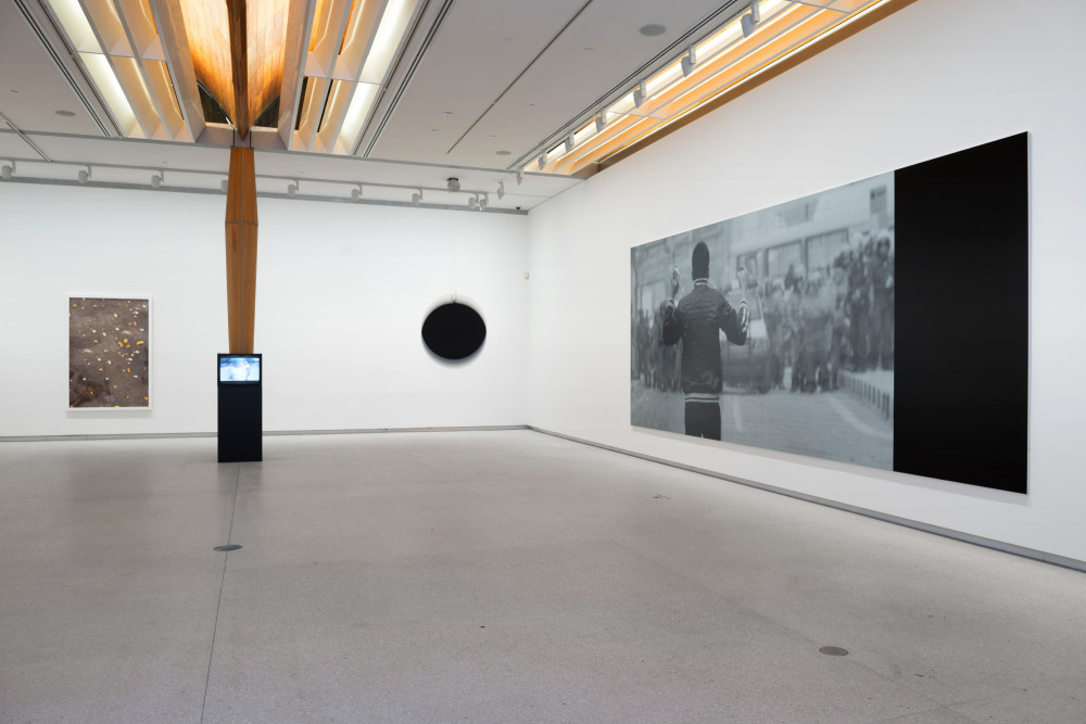 The image shows the corner of a gallery room. On the left hand wall, far left, is a tall framed photograph depicting orange peels scattered across dark grey tarmac. To the right of the photograph in front of a tall wooden column is a small cube television set on a plinth, playing a blue tinted film. To the right of this in the corner is a large black sphere attached to string, which is moving in an agitated manner. On the right hand wall hangs a large horizontal photograph in black and white of a man with his back to the viewer, facing a crowd of people and a stationary car in a street. 