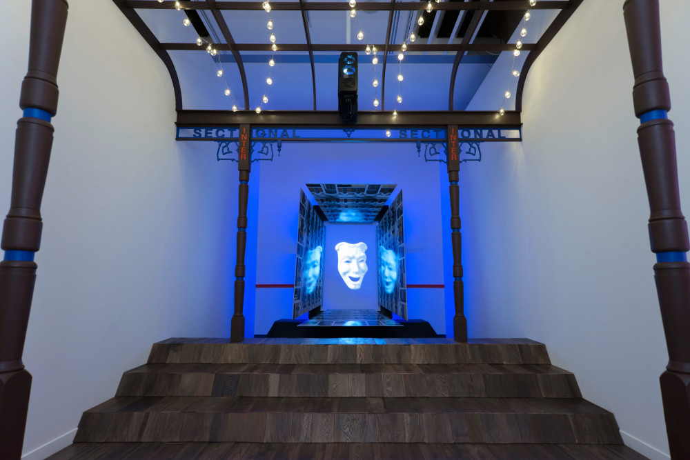 The image shows a small enclosed gallery space. A large metal structure fills the space, with thin columns rising to the ceiling and a grid-like metal roof with the words 'sectional' running along the edges of the roof on each side. A flight of large dark stairs lead the viewer up to a hologram of a smiling comedy mask which floats in a rectangular recess, with blue light surrounding it. 